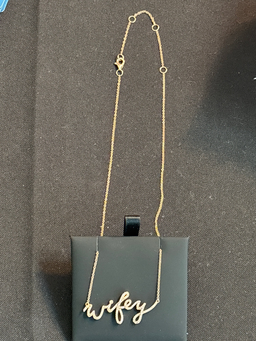 A gold script necklace spelling 'wifey' draped over a black display card on a dark textured background.