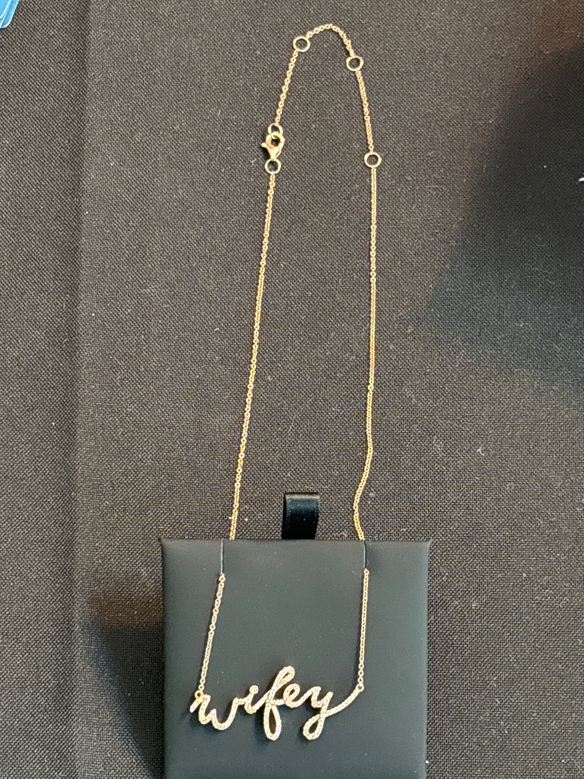 A gold script necklace spelling 'wifey' draped over a black display card on a dark textured background.