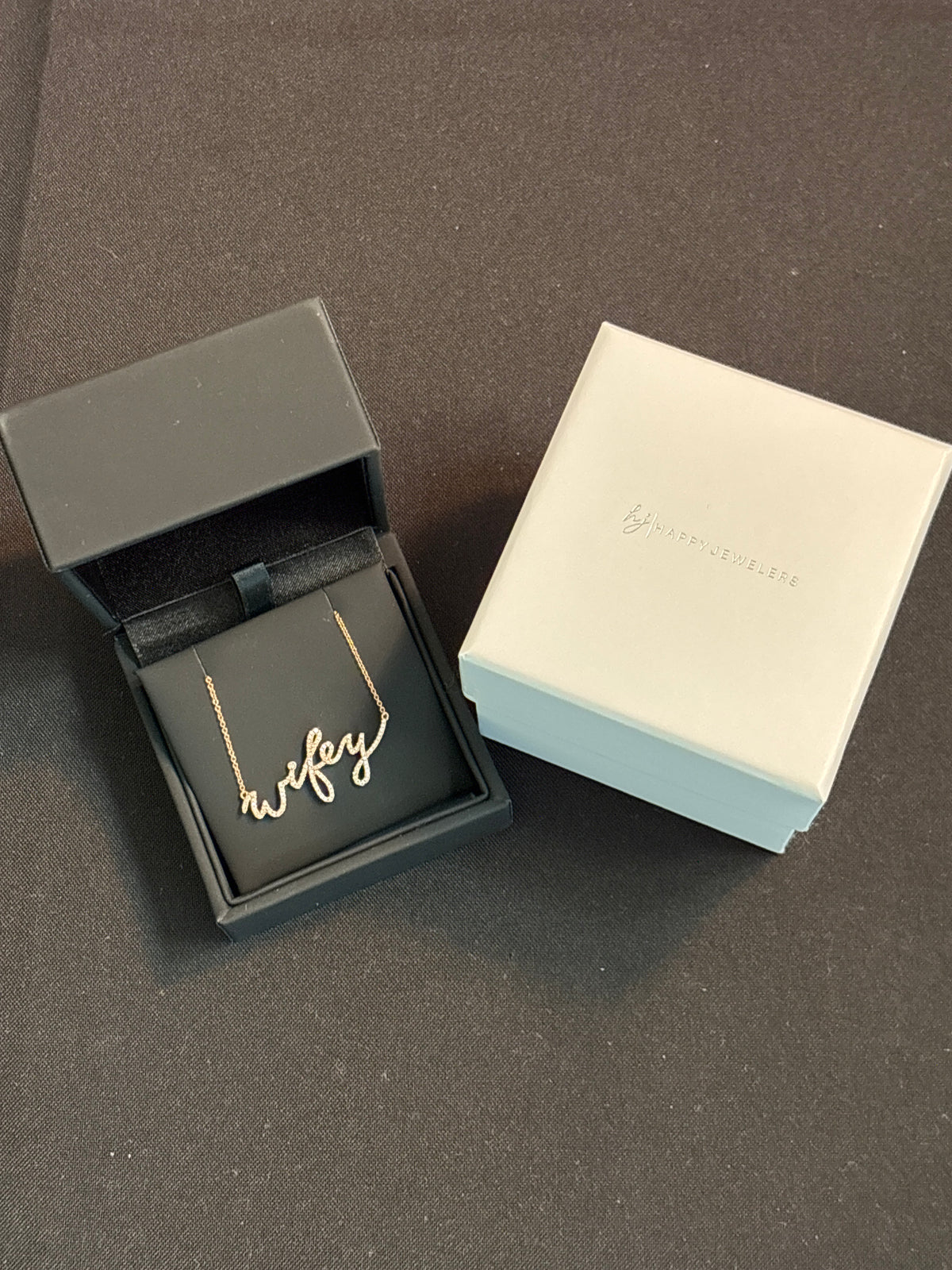A gold script name necklace reading 'wifey” is displayed inside an open black jewelry box next to its white branded packaging box on a dark gray surface.